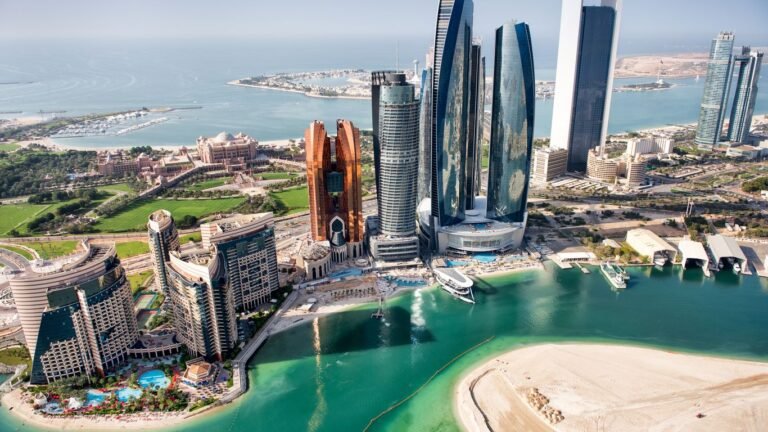Aerial view of Abu Dhabi waterfront skyline with luxury towers, turquoise water, marina, and coastal landmarks.